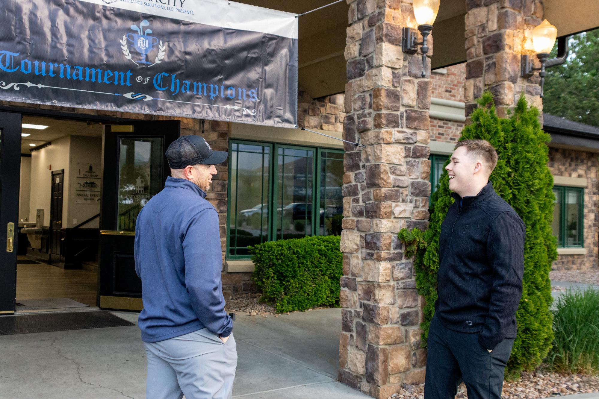 Two golfers chatting and laughing under the Tournament of Champions sign at the clubhouse entrance