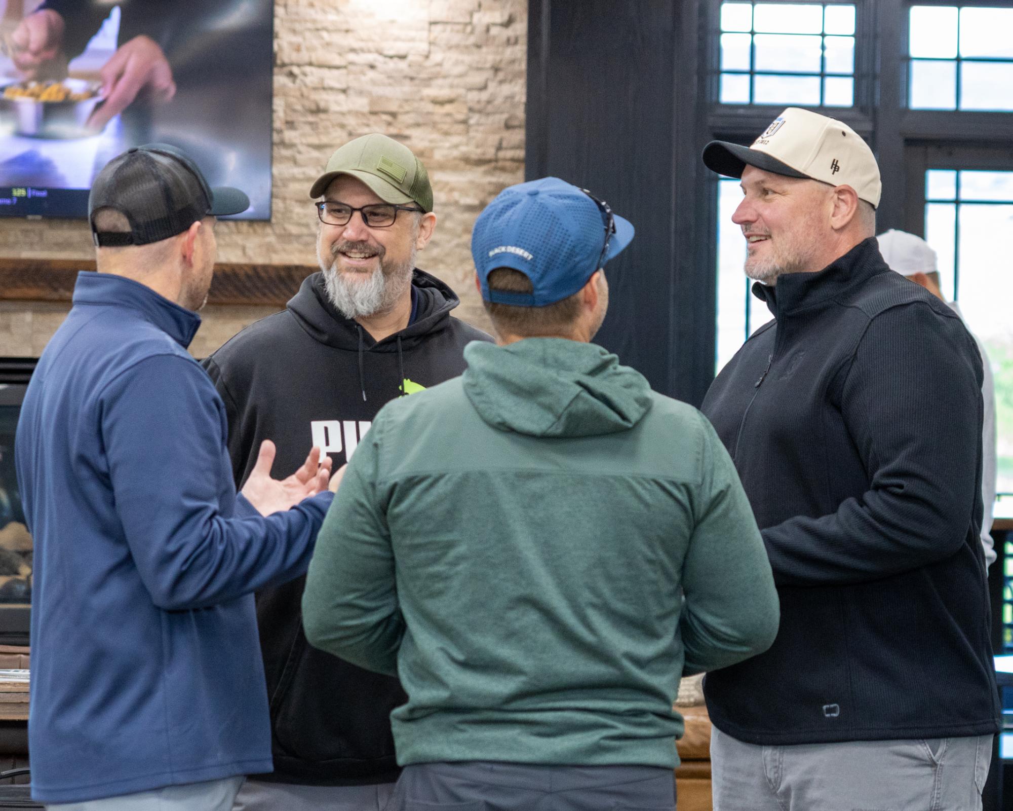 Small group of golfers standing and talking in the clubhouse near a stone fireplace and TV screens
