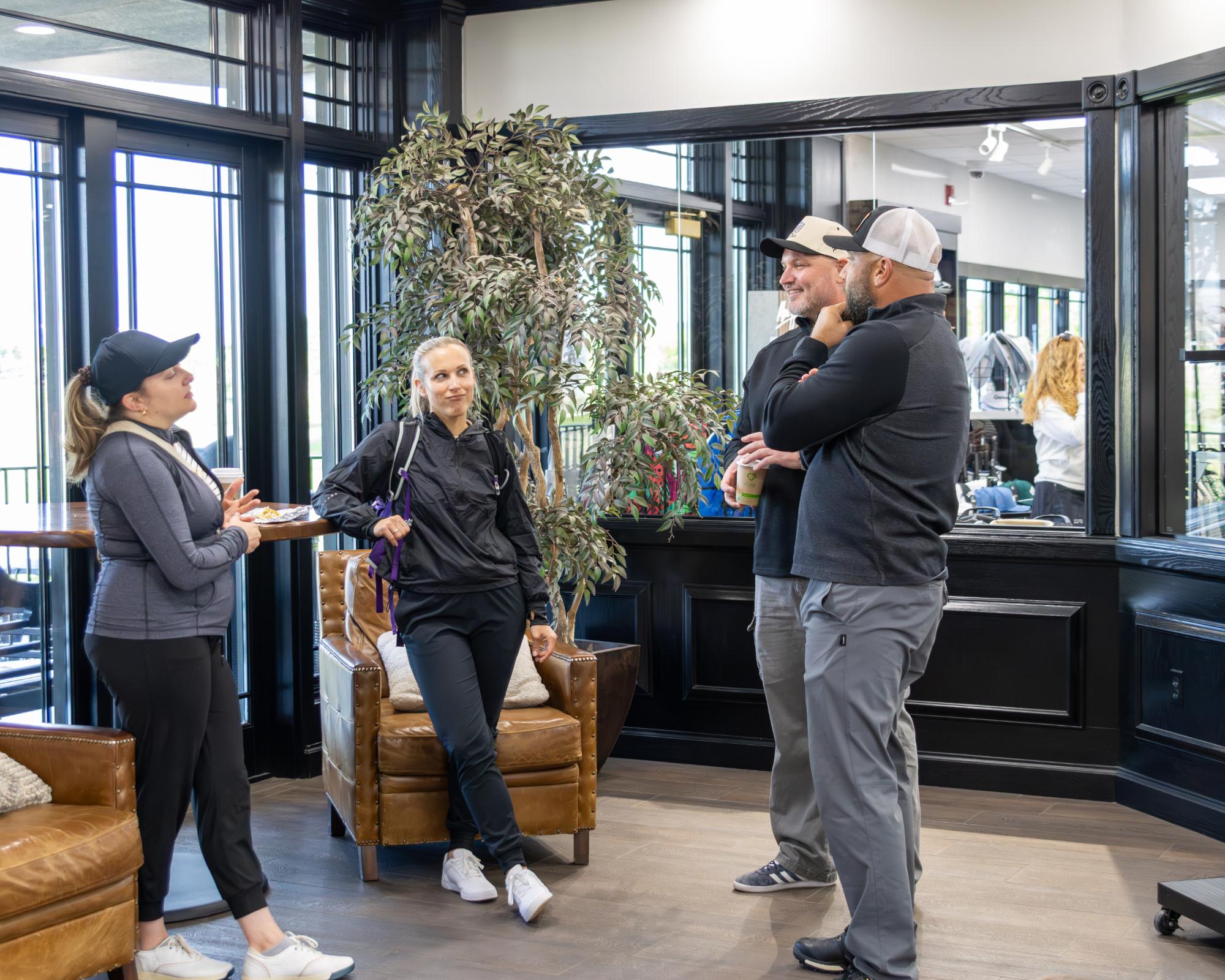 Three players in casual golf attire chatting in a lounge area with large indoor windows