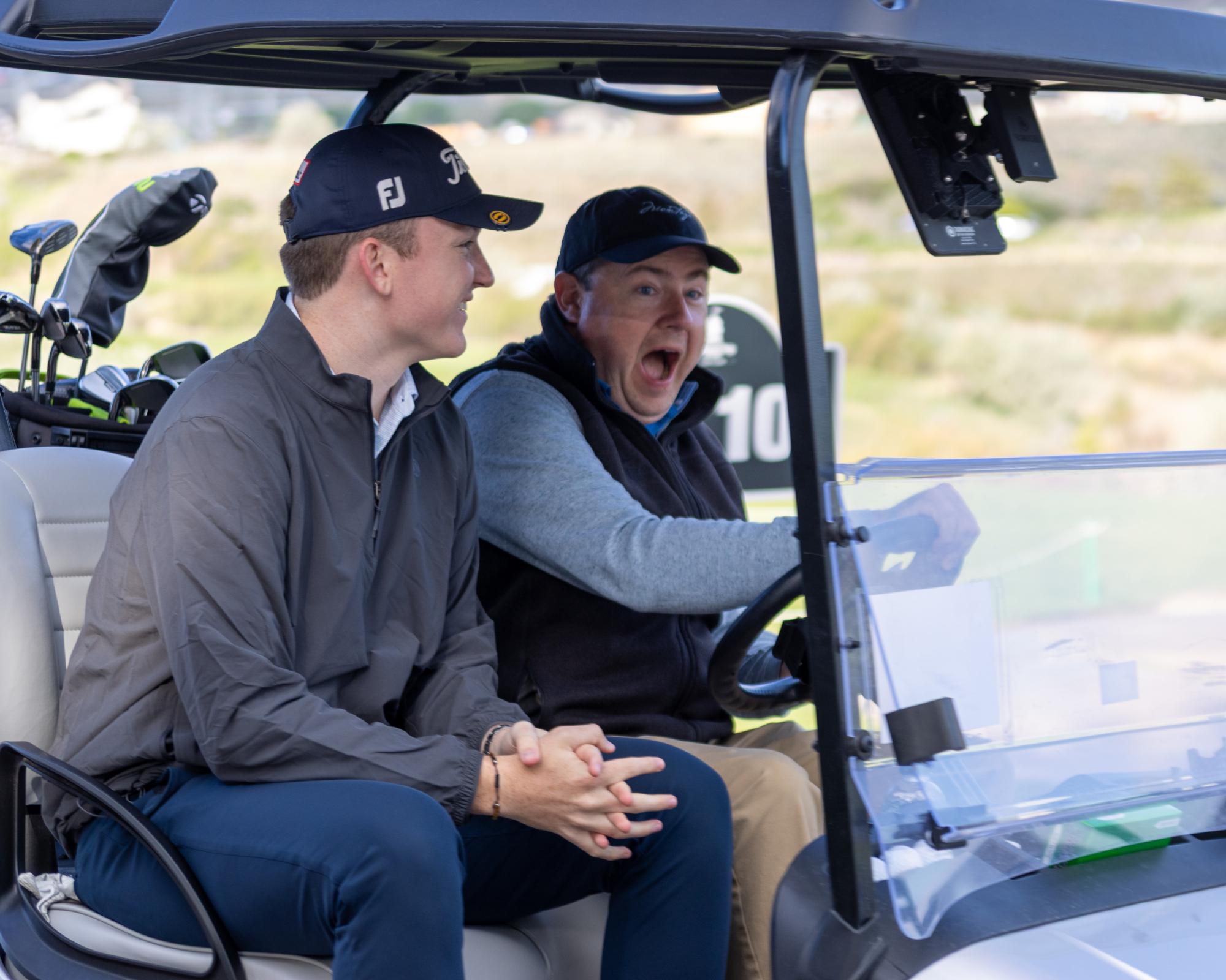 Two friends seated in a golf cart, one yawning playfully while the other smiles ahead