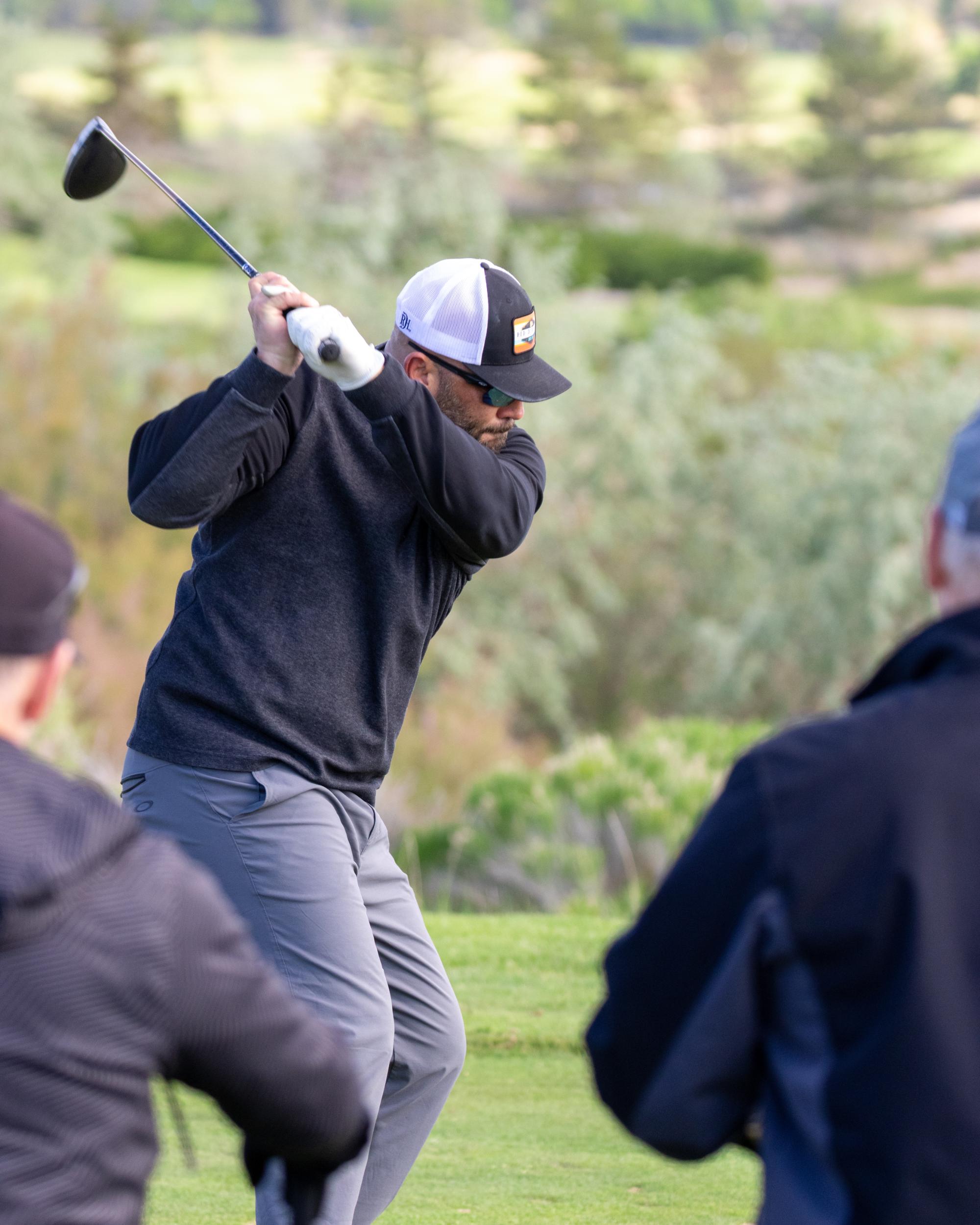 Golfer in black long-sleeve shirt and white cap wearing sunglasses executing a full driver swing at the tee