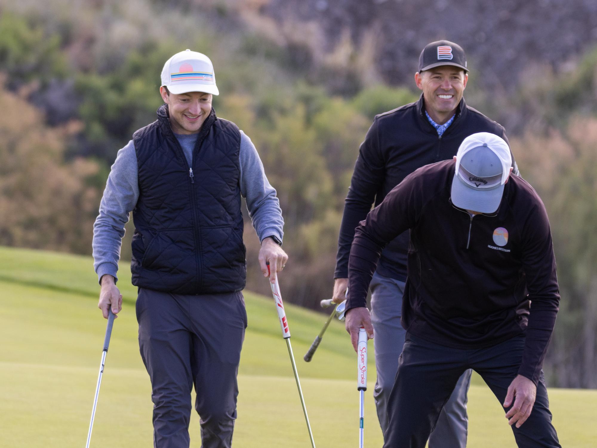 Three golfers walking together on the putting green, chatting and carrying their putters after a hole