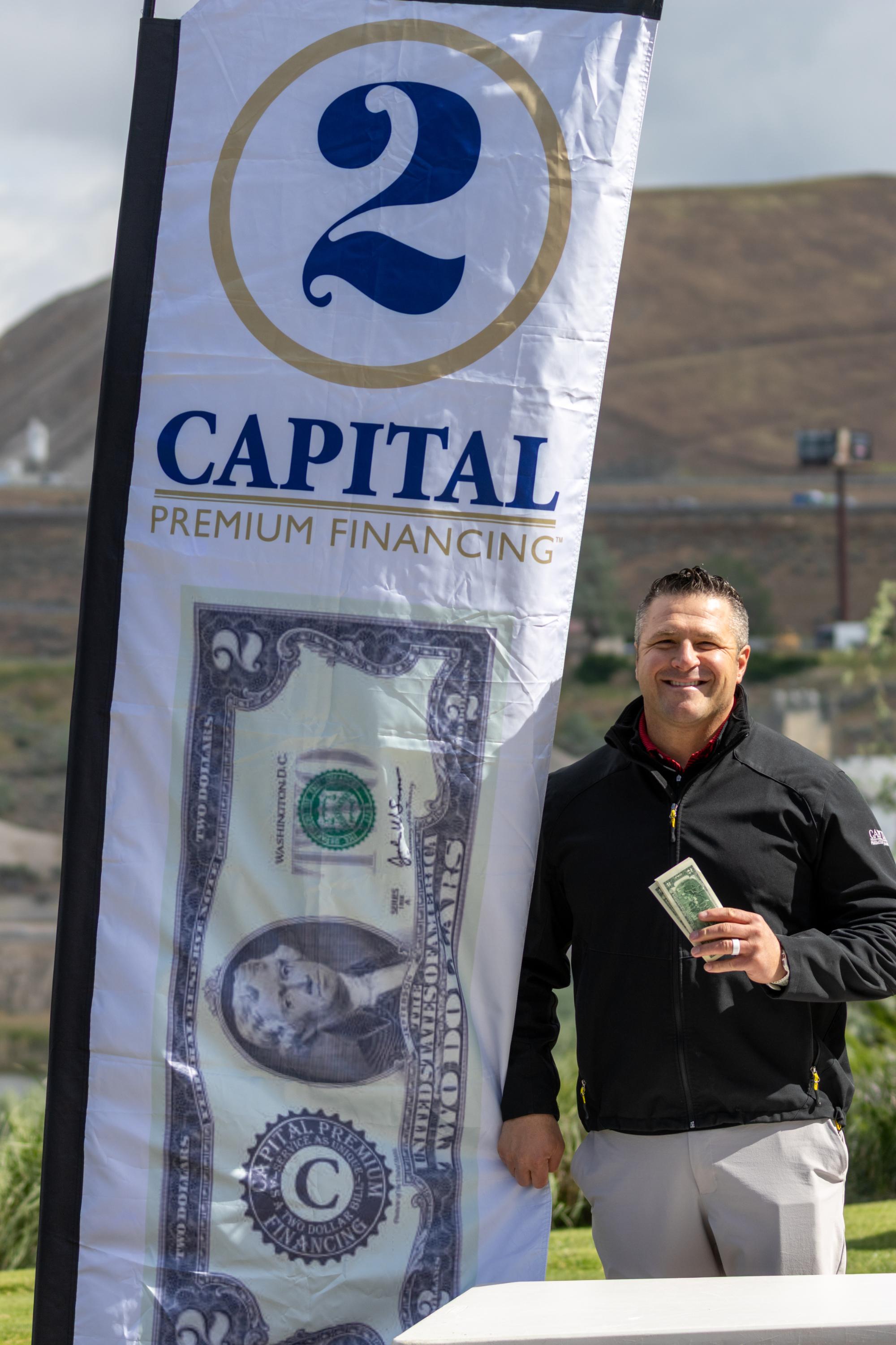 Participant standing next to a Capital Premium Financing flag with an oversized two-dollar bill banner, holding cash and smiling