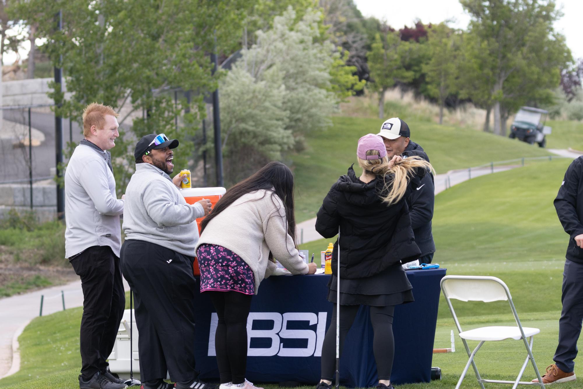 Golfers checking in at a BSI registration table on the fairway, laughing and signing scorecards