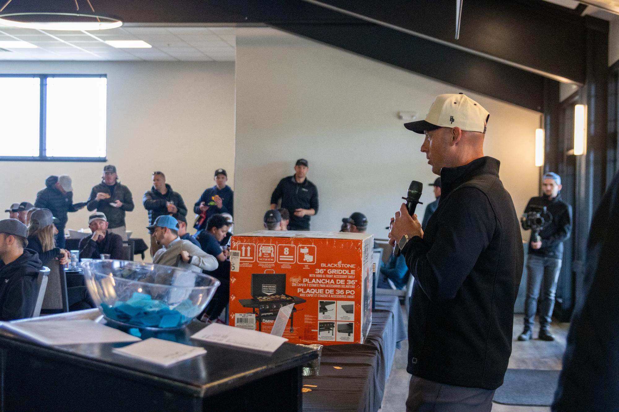 Tournament organizer speaking into a microphone next to a boxed griddle prize with seated audience in background